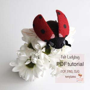 May include: A handmade felt ladybug with red wings and black spots, perched on a cluster of white flowers. The ladybug has a black head and body. The image includes the text "Felt Ladybug PDF tutorial".
