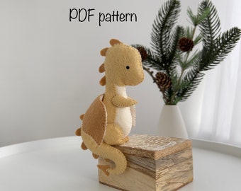May include: A felt dragon toy with a light brown body, a tan back, and a white belly. The dragon is sitting on a wooden box with the text "PDF pattern" above it.