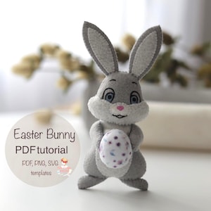 Puede incluir: Un conejito de Pascua de fieltro gris sosteniendo un huevo decorado. El conejito tiene orejas largas, ojos azules y una nariz rosa. La imagen incluye el texto "Easter Bunny PDF tutorial" y "PDF, PNG, SVG templates".