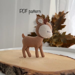 May include: A brown felt deer with white felt ears and a brown felt nose. The deer is standing on a wooden surface. The text "PDF pattern" is written above the deer.