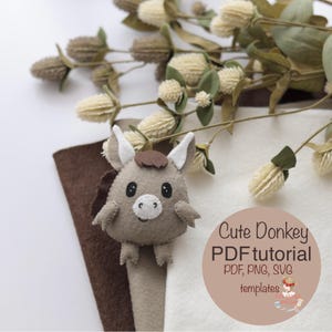 May include: A handmade felt donkey bookmark with a brown mane and ears, and a white muzzle. The bookmark is light gray and brown, with the text "Cute Donkey PDF tutorial" in a circular design. Dried flowers are in the background.