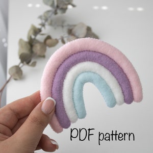 Peut inclure: Un patron PDF pour un artisanat en feutre en forme d'arc-en-ciel. L'arc-en-ciel est composé de quatre couches de feutre en rose, violet, blanc et bleu clair.