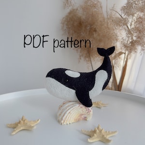 May include: PDF pattern for a felt orca whale toy. The orca is black and white with a white belly and a black back. The orca is sitting on a seashell.