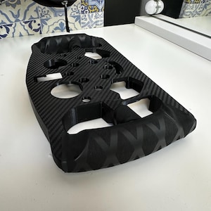 May include: Black carbon fiber steering wheel with multiple circular cutouts and a textured grip. The wheel has a distinctive pattern and is designed for a vehicle. The background includes a white surface and a decorative tile pattern.