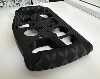 Puede incluir: Volante negro de fibra de carbono con múltiples recortes circulares y un agarre texturizado. El volante tiene un patrón distintivo y está diseñado para un vehículo. El fondo incluye una superficie blanca y un patrón de baldosas decorativas.