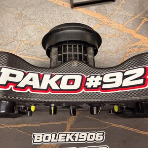 May include: A black and gray automotive part with the text "PAKO #92" in white and red. The part has a carbon fiber pattern and several black and yellow knobs. The text "BOLEK1906" is also visible.