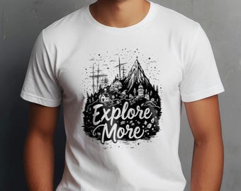 May include: White t-shirt featuring a black and white graphic of a mountain, buildings, and trees. The text "Explore More" is written in a stylized font below the graphic. The shirt has short sleeves and a crew neck.