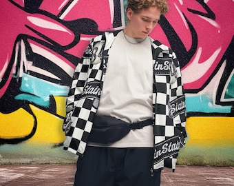 Op de afbeelding: Een persoon draagt een zwart-wit geruit jack met het woord "Stain" erop gedrukt, een wit t-shirt, een zwarte broek en een zwart heuptasje. De achtergrond toont kleurrijke graffiti-kunst.