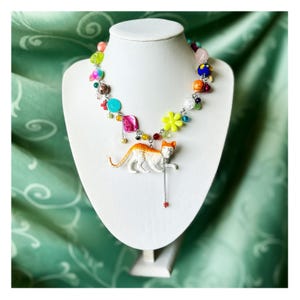 May include: A colorful beaded necklace with a cat pendant. The necklace features a variety of beads in shades of pink, blue, yellow, and green. The cat pendant is white with orange markings and hangs from a silver chain.