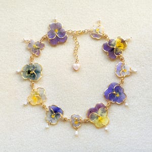 Puede incluir: Un delicado collar con flores de pensamiento prensadas en morado, amarillo y blanco. Las flores están engarzadas en metal dorado con pequeños detalles de perlas. El collar tiene un dije en forma de corazón, sobre fondo blanco.