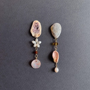 May include: A pair of handcrafted earrings with natural seashell elements. One earring includes a white flower charm, red bead, and a pink iridescent disc. The other features a pink shell, a pearl, and gold-coloured accents. Set against a grey backdrop.