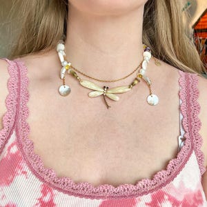 Puede incluir: Un collar en capas con un colgante de libélula. El collar presenta una cadena dorada, cuentas de concha blancas y pequeños dijes de disco dorados. El colgante de libélula es de color crema con detalles en negro.