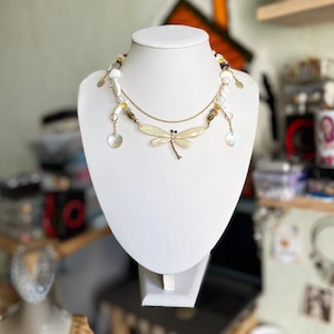 Puede incluir: Un collar artesanal con un colgante de libélula y una mezcla de cuentas blancas, amarillas y marrones. El collar incluye colgantes en forma de concha y se exhibe en un busto de maniquí blanco. El colgante de libélula es de color crema con detalles dorados.