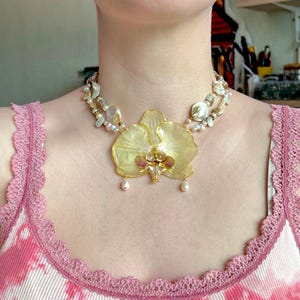 Könnte beinhalten: Eine handgefertigte Halskette mit einem großen, gelben Orchideenanhänger, goldfarbenen Details und zwei hängenden Perlen. Die Halskette hat zwei Stränge aus weißen Perlen. Die Person trägt ein rosa-weißes Oberteil.