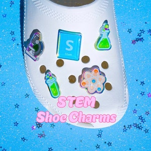 May include: A set of five iridescent STEM shoe charms. The charms are shaped like a microscope, a beaker, a molecule, a letter S, and a test tube. The charms are on a white shoe with a blue background.