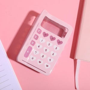 Puede incluir: Una calculadora rosa con botones en forma de corazón. La calculadora tiene una ventana de visualización clara y una carcasa de plástico rosa.