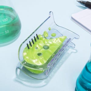 以下が含まれることがあります： 明るい緑色のテクスチャのある物質で満たされた、ビーカー型の透明なプラスチッククリップ。 クリップには黒い線があり、他の実験器具が見える淡い青色の背景に配置されています。