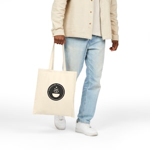 May include: Cream-colored tote bag with a black circular logo featuring a coffee bean and leaf design. The bag is held by a person wearing light-wash jeans, white sneakers, and a beige button-up shirt. The text "CAFETO 144" is above the logo.