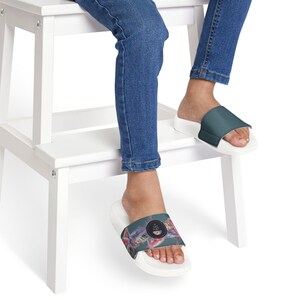 Puede incluir: Sandalias deslizantes blancas con una correa verde azulado y un diseño abstracto colorido. Las sandalias las lleva una persona con vaqueros azules. Las sandalias tienen suela blanca y correa verde azulado.