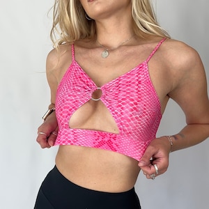 Könnte beinhalten: Ein hot pinkes Crop-Top mit Schlangenmuster, Schlüsselloch-Design und silbernem Ringdetail. Kombiniert mit schwarzen Sporthosen. Das Top hat dünne Spaghettiträger.