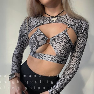 Könnte beinhalten: Ein schwarzes und weißes Schlangenmuster-Crop-Top mit langen Ärmeln, einem Ausschnitt und Metallringen. Das Oberteil hat eine figurbetonte Silhouette.