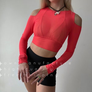 Könnte beinhalten: Ein leuchtend rotes, langärmliges Crop-Top mit ausgeschnittenen Schultern. Das Oberteil ist aus einem durchsichtigen, leichten Stoff gefertigt. Das Model trägt eine schwarze Shorts.