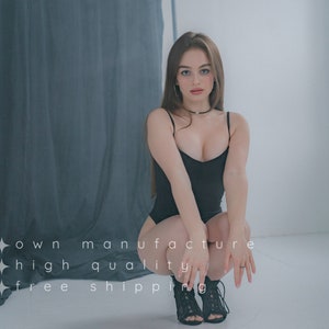 May include: A woman wearing a black sleeveless bodysuit and black heeled shoes poses in front of a gray backdrop. She is looking at the camera with a serious expression.