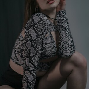 K&ouml;nnte beinhalten: Eine Frau tr&auml;gt ein Oberteil mit Schlangenmuster in Schwarz und Wei&szlig; mit einem schwarzen Crop-Top darunter. Sie hockt mit der Hand an der Wange und blickt in die Kamera.