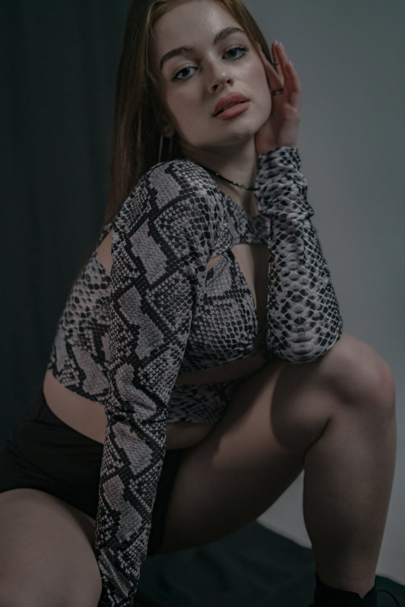 K&ouml;nnte beinhalten: Eine Frau tr&auml;gt ein Oberteil mit Schlangenmuster in Schwarz und Wei&szlig; mit einem schwarzen Crop-Top darunter. Sie hockt mit der Hand an der Wange und blickt in die Kamera.