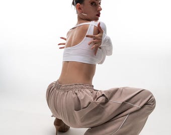 Könnte beinhalten: Ein weißes Crop-Top mit langen Ärmeln und Cut-Out-Schultern und eine beige Hose mit weißem Besatz. Das Model trägt High Heels. Das Outfit ist ein moderner, minimalistischer Stil.