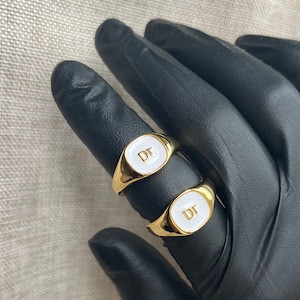 May include: Two gold-colored signet rings with white enamel faces, each displaying the letters "DT" in gold. The rings are oval-shaped and rest on a black surface.