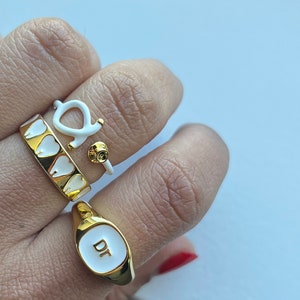 May include: A hand displays four gold rings with white enamel accents. One ring features a row of white teeth, another has a white stethoscope design, and a third has a small gold and white design. The fourth ring is gold with the letters "DT" on a white background.