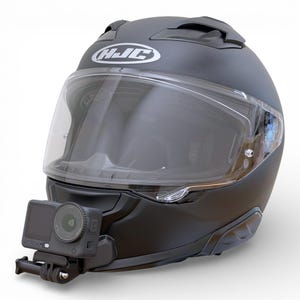 Könnte beinhalten: Mattschwarzer Motorradhelm mit klarem Visier und weißem "HJC"-Logo. Eine Action-Kamera ist vorne am Helm befestigt. Die Kamera ist schwarz und hat ein Objektiv und einen Bildschirm.