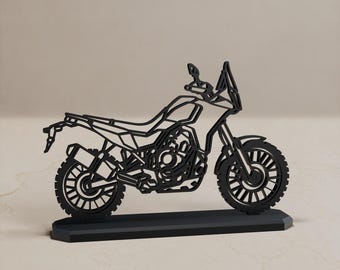 Könnte beinhalten: Eine schwarze, flache, lasergeschnittene Silhouette eines Abenteuermotorrads auf einem rechteckigen Sockel. Das Design zeigt eine offene Rahmenstruktur mit Rädern, Motor, Lenker und Windschutzscheibe in einer stilisierten, linearen, zweidimensionalen Form.