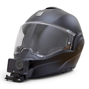 Peut inclure: Un casque de moto modulaire noir mat doté d'une visière transparente et d'une petite caméra d'action noire montée sur la mentonnière. Le casque présente une coque lisse et arrondie avec un emblème argenté situé au centre supérieur.