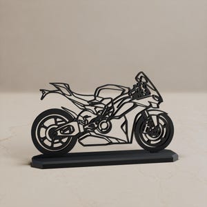 Może przedstawiać: Czarna, płaska, wycinana laserowo metalowa sylwetka motocykla sportowego osadzona na prostokątnej podstawie. Projekt charakteryzuje się otwartą konstrukcją ramową, zarysowującą koła, silnik, owiewki oraz kierownicę motocykla w jednej linii.