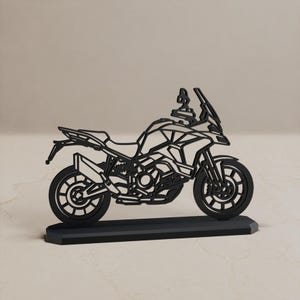Może przedstawiać: Czarna, płaska, wycinana laserowo metalowa sylwetka motocykla typu adventure, umieszczona na prostokątnej podstawie. Projekt charakteryzuje się ażurową konstrukcją przedstawiającą koła, silnik, wydech, kierownicę oraz szybę pojazdu.