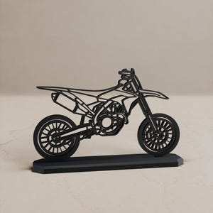 Może przedstawiać: Czarna, płaska, wycinana laserowo sylwetka motocykla motocrossowego osadzona na prostokątnej podstawie. Projekt posiada otwartą konstrukcję ramową przedstawiającą koła, silnik, siedzenie, kierownicę oraz rurę wydechową motocykla typu dirt bike.