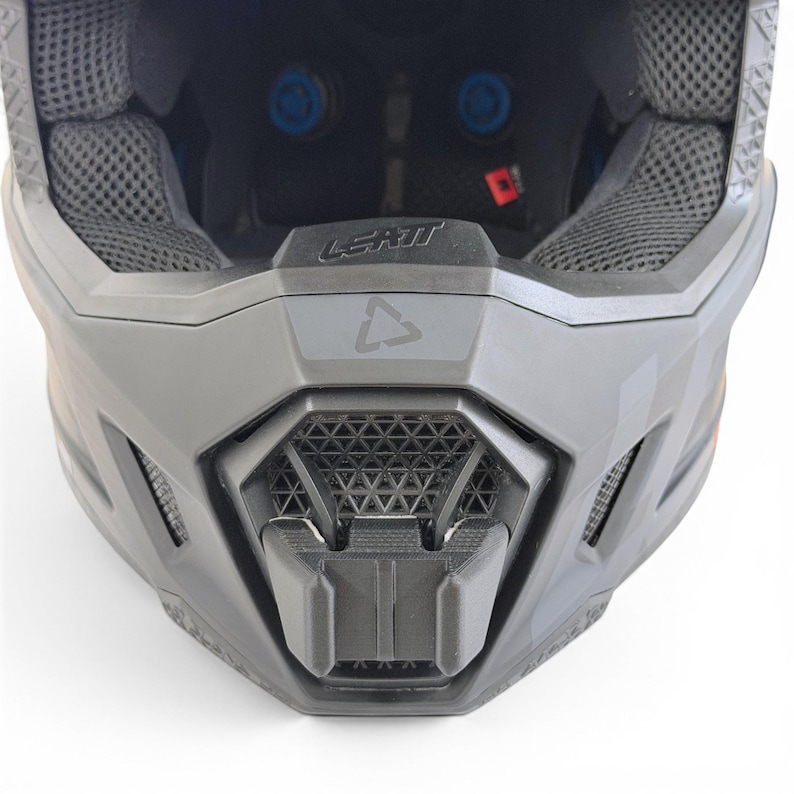 May include: A close-up view of a gray motocross helmet. The helmet features a geometric design on the chin guard and the brand name "LEATT" is visible. The interior padding is gray and the helmet has blue accents.
