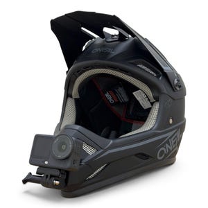 May include: A matte black O'Neal full-face motocross helmet featuring a large adjustable visor, ventilation ports with mesh inserts, and a black action camera mounted securely to the chin bar. The interior padding is light grey with red accents.