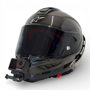 Puede incluir: Un casco de moto negro brillante con una visera oscura y una cámara de acción montada. El casco presenta un patrón de tejido de fibra de carbono y logotipos de estrellas blancas. Un acento rojo es visible cerca de la barbilla.
