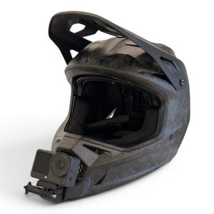 May include: A black and grey Fox full-face helmet with a visor and an attached action camera. The helmet has a textured pattern and is designed for sports activities. The camera is mounted on the chin area.