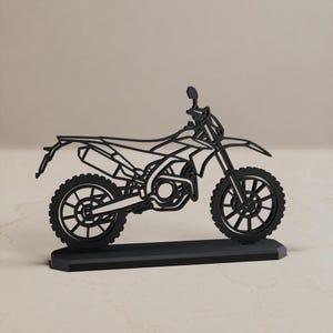 Może przedstawiać: Czarna, płaska, wycinana laserowo sylwetka motocykla typu dirt bike umieszczona na prostokątnej podstawie. Projekt charakteryzuje się otwartą konstrukcją ramy, ukazującą koła, silnik, siedzenie, kierownicę oraz rurę wydechową w profilu.