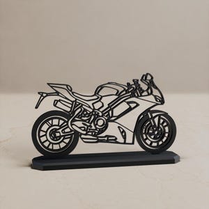 Może przedstawiać: Czarna, płaska, wycinana laserowo sylwetka motocykla sportowego umieszczona na prostokątnej podstawie. Projekt przedstawia szczegółowe zarysy kół, silnika, ramy, siedzenia i kierownicy, tworząc ażurową, otwartą strukturę pojazdu.
