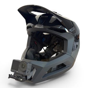 May include: A black full-face mountain bike helmet with a matte and glossy finish, featuring a prominent visor and the Leatt brand logo. A grey action camera is securely mounted to the chin bar, positioned to face forward for recording.