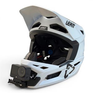 May include: A white full-face mountain bike helmet with black accents and the Leatt logo printed on the side and front. A black action camera is mounted to the chin bar. The helmet features multiple ventilation ports and a large adjustable visor.