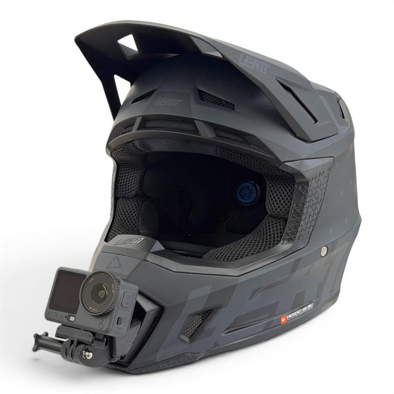 May include: A matte black LEATT full-face helmet with a visor and integrated camera mount. The helmet has a star pattern and mesh ventilation. A black action camera is mounted on the front.
