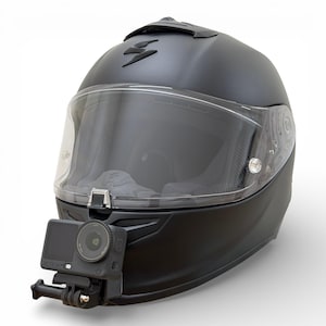 Puede incluir: Casco de moto negro mate con visera transparente y cámara de acción montada. El casco presenta un logotipo de Scorpion y un diseño aerodinámico, ideal para motociclistas.