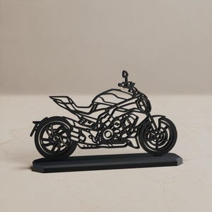 Może przedstawiać: Czarna, płaska, wycinana laserowo sylwetka motocykla typu naked, osadzona na prostokątnej podstawie. Projekt zawiera skomplikowane wycięcia przedstawiające silnik, ramę, koła i kierownicę, tworząc szczegółowy profil z ażurowymi detalami.
