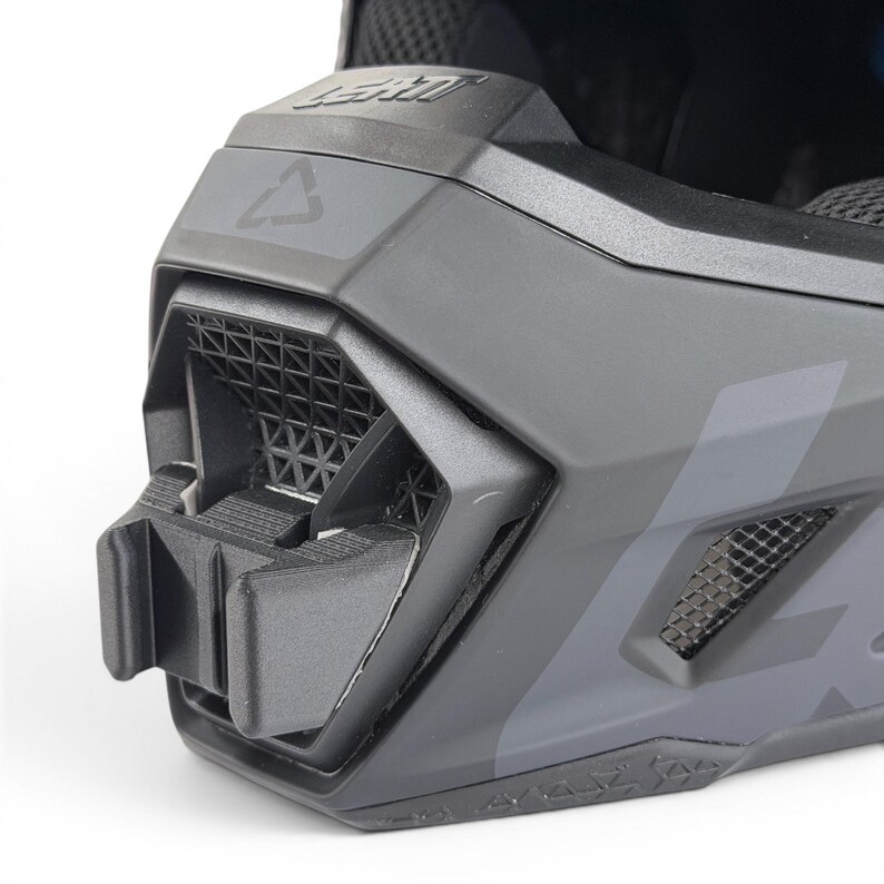 May include: Close-up of a matte gray LEATT brand helmet. The helmet features a geometric design with ventilation grilles and a mounting bracket. The LEATT logo is visible on the top. The helmet is designed for protection and performance.